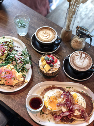 A top down view of breakfast. Smoked salmon and scrambled eggs on sourdough toast, bacon and egg pancakes, a coconut smoothie bowl and flat white coffees